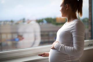 Pregnant woman in a white top stands by a window, hand on her belly, looking outside.